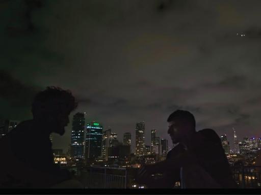 Zwei Männer sind im Profil zu erkennen. Sie sitzen im Dunkeln draußen. Im Hintergrund ist die Skyline von Tel Aviv zu erkennen. Es handelt sich um eine Szene aus der Doku "NAZA".