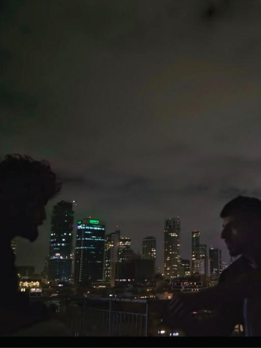 Zwei Männer sind im Profil zu erkennen. Sie sitzen im Dunkeln draußen. Im Hintergrund ist die Skyline von Tel Aviv zu erkennen. Es handelt sich um eine Szene aus der Doku "NAZA".