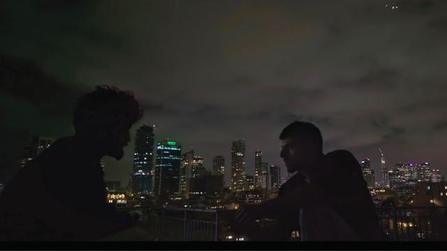 Zwei Männer sind im Profil zu erkennen. Sie sitzen im Dunkeln draußen. Im Hintergrund ist die Skyline von Tel Aviv zu erkennen. Es handelt sich um eine Szene aus der Doku "NAZA".