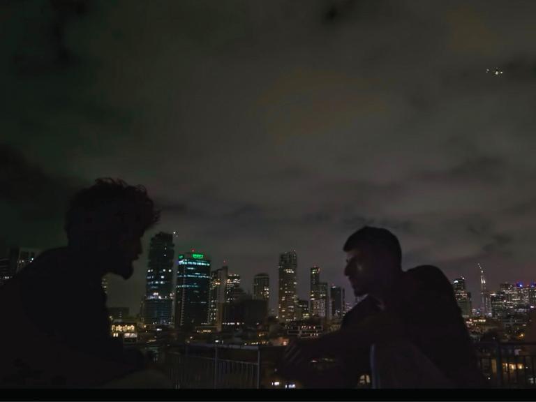 Zwei Männer sind im Profil zu erkennen. Sie sitzen im Dunkeln draußen. Im Hintergrund ist die Skyline von Tel Aviv zu erkennen. Es handelt sich um eine Szene aus der Doku "NAZA".