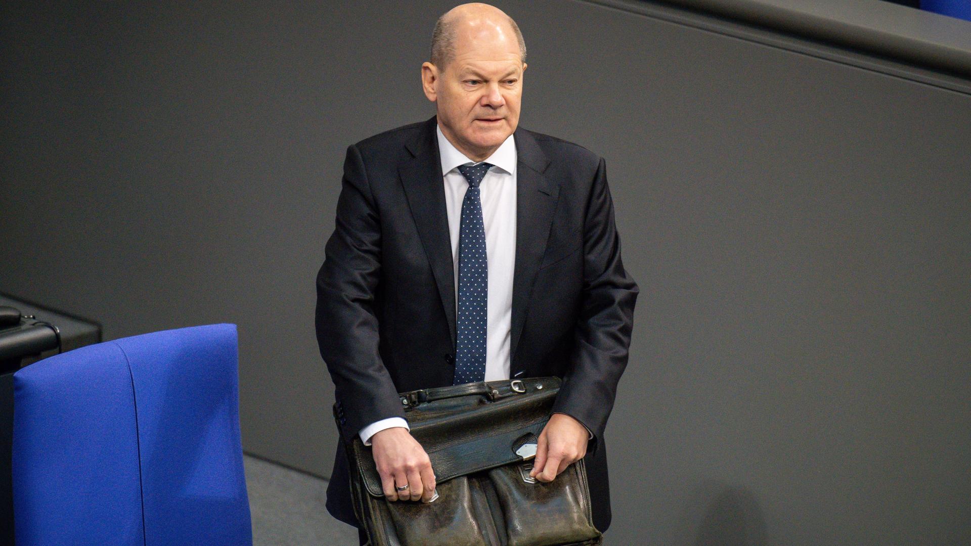 Bundeskanzler Olaf Scholz steht mit einer ledernen Aktentasche an einer der Stuhlreihen im Deutschen Bundestag.