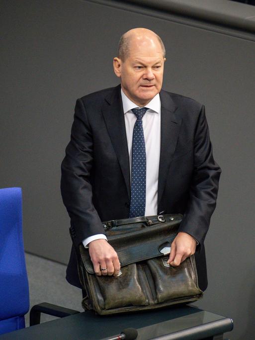 Bundeskanzler Olaf Scholz steht mit einer ledernen Aktentasche an einer der Stuhlreihen im Deutschen Bundestag. Bundeskanzler Olaf Scholz steht mit einer ledernen Aktentasche an einer der Stuhlreihen im Deutschen Bundestag.