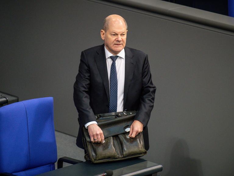 Bundeskanzler Olaf Scholz steht mit einer ledernen Aktentasche an einer der Stuhlreihen im Deutschen Bundestag. Bundeskanzler Olaf Scholz steht mit einer ledernen Aktentasche an einer der Stuhlreihen im Deutschen Bundestag.
