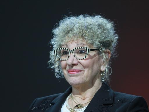 Ein Foto der Schriftstellerin Lizzie Doron aufgenommen am 21.10.2023 auf der Frankfurter Buchmesse. Sie sitzt vor einem schwarzen Hintergrund, wahrscheinlich einer Bühne, hat ein leichtes Lächeln auf den Lippen und trägt eine auffällige Brille.