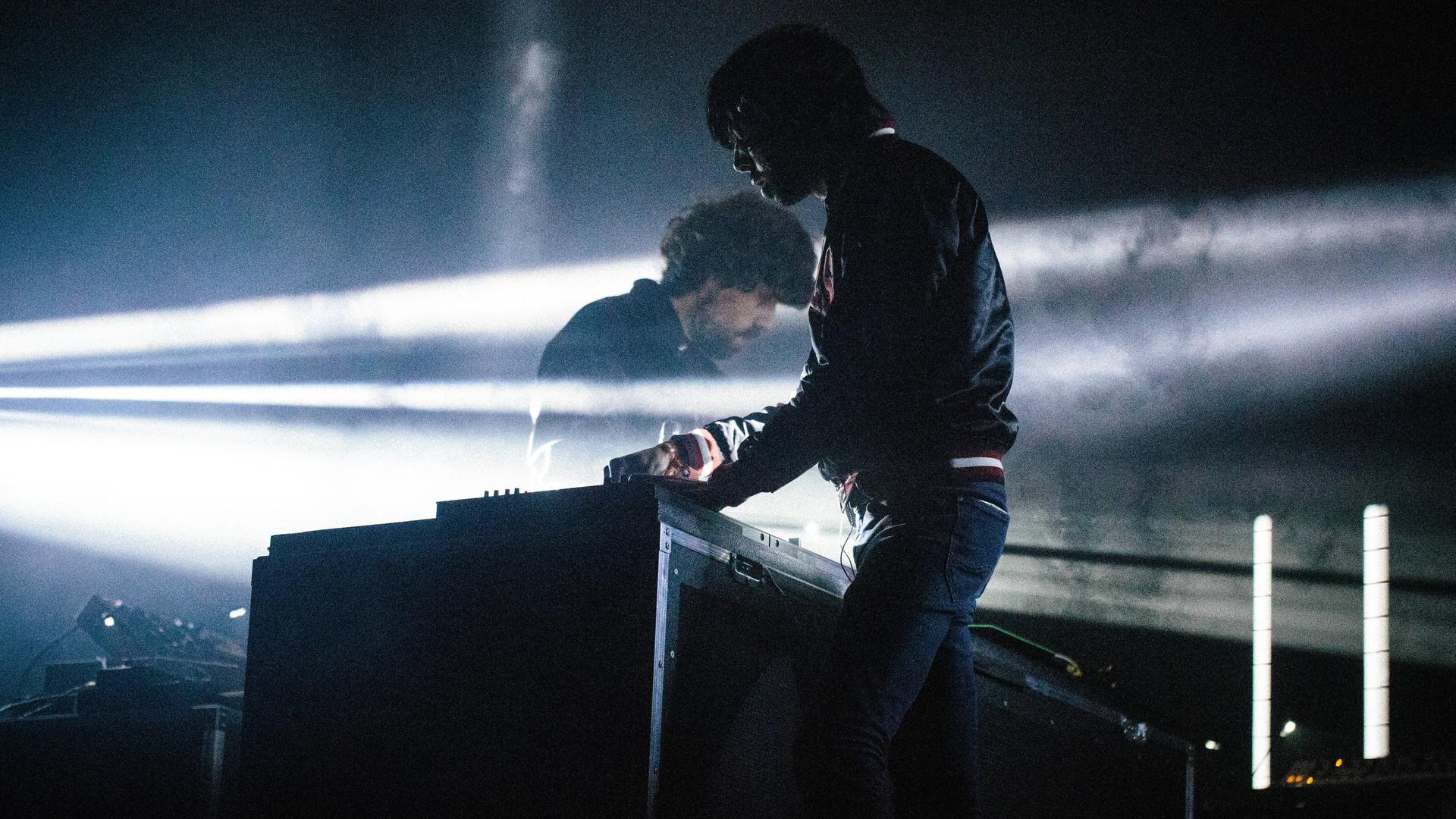 Die zwei Mitglieder von Justice stehen während eines Auftrittes auf einer dunklen Bühne im Lichtschein.