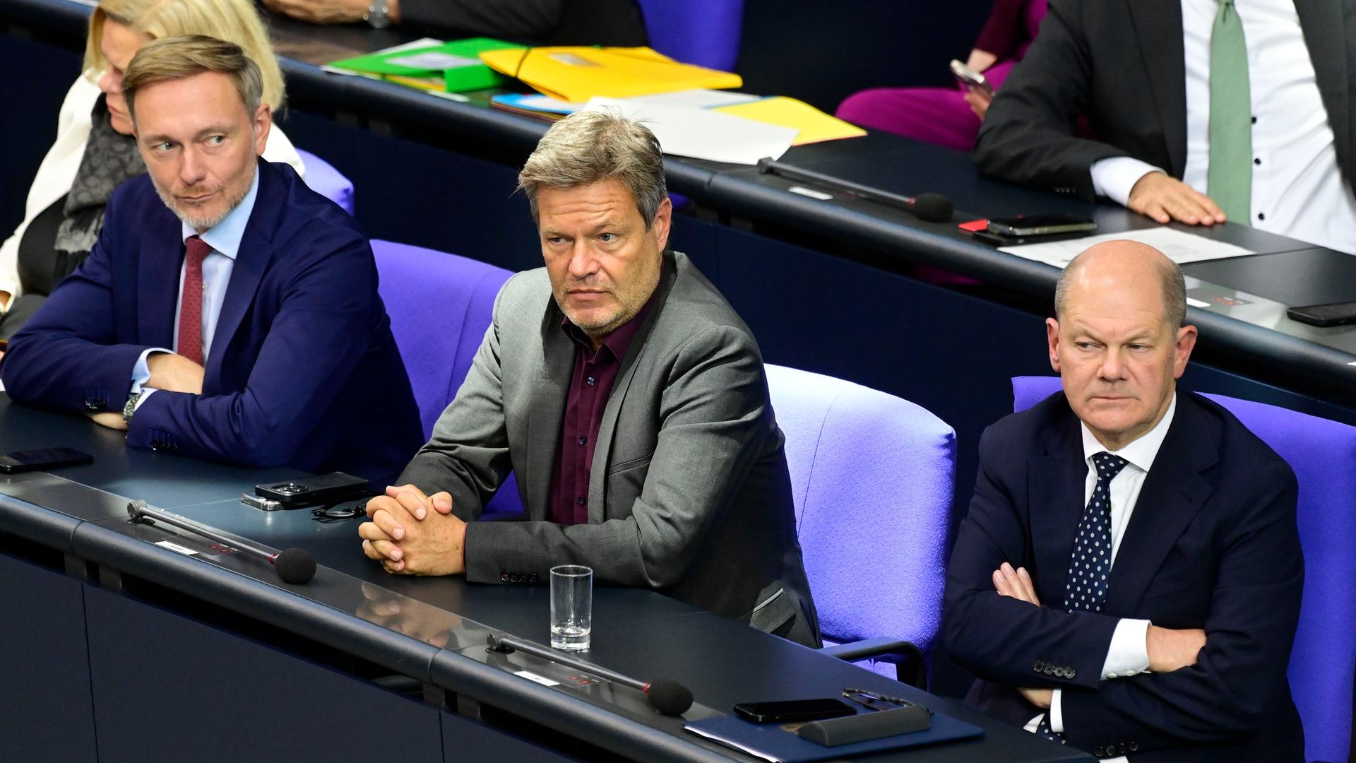 Christian Lindner, Robert Habeck und Olaf Scholz sitzen nebeneinander auf ihren Plätzen im Bundestag. Lindner schielt zu Habeck und Scholz. Habeck und Scholz schauen grimmig nach vorne.