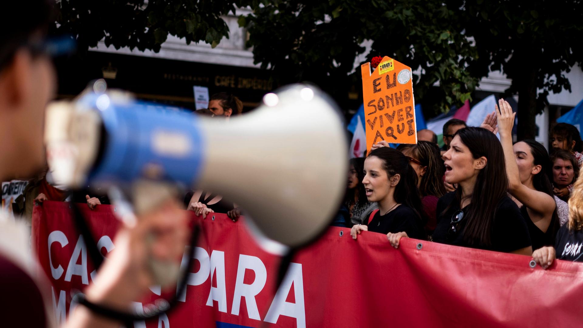 Zahlreiche Menschen laufen bei einer Demonstration mit Transparenten durch die Straßen; im Vordergrund ein unscharfes Megafon.