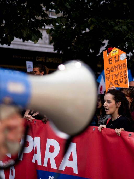Zahlreiche Menschen laufen bei einer Demonstration mit Transparenten durch die Straßen; im Vordergrund ein unscharfes Megafon. Zahlreiche Menschen laufen bei einer Demonstration mit Transparenten durch die Straßen; im Vordergrund ein unscharfes Megafon.