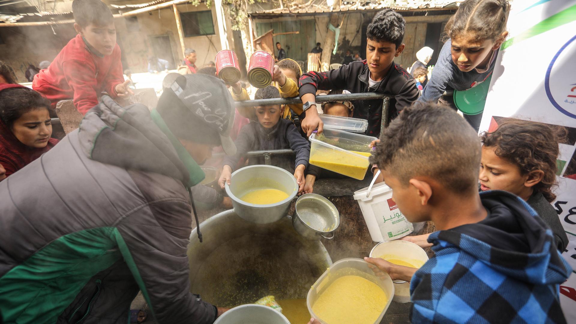 Im Gazastreifen stehen Kinder um Essen an, sie halten Behälter in den Händen. In der Mitte des Fotos steht ein Topf, daraus verteilt eine Person Suppe.