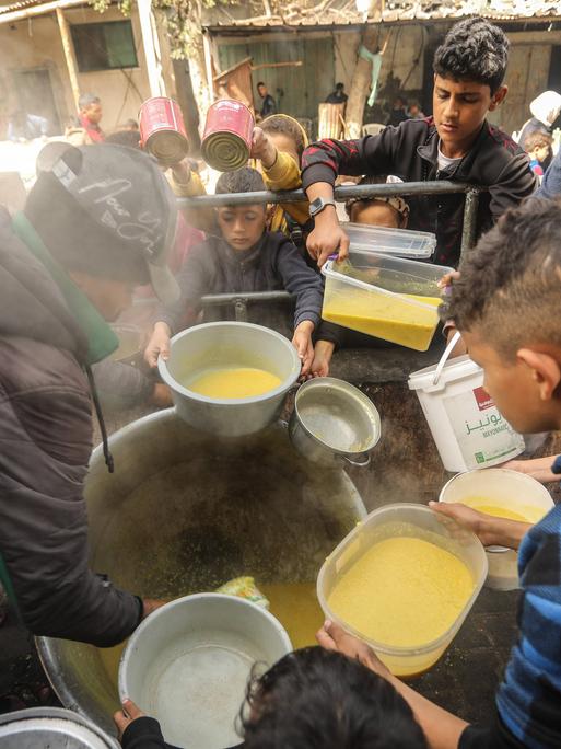 Im Gazastreifen stehen Kinder um Essen an, sie halten Behälter in den Händen. In der Mitte des Fotos steht ein Topf, daraus verteilt eine Person Suppe. (6.3.2024)