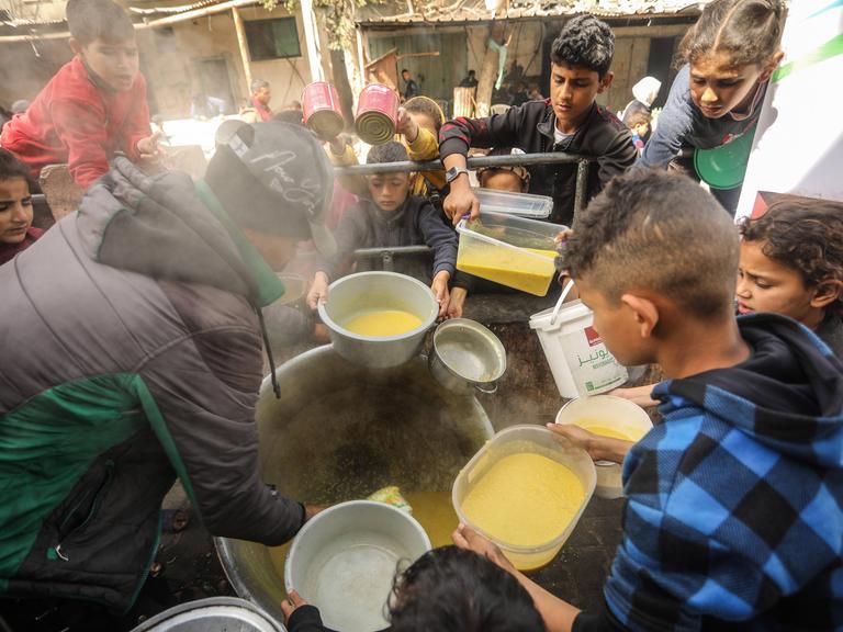 Im Gazastreifen stehen Kinder um Essen an, sie halten Behälter in den Händen. In der Mitte des Fotos steht ein Topf, daraus verteilt eine Person Suppe. (6.3.2024)