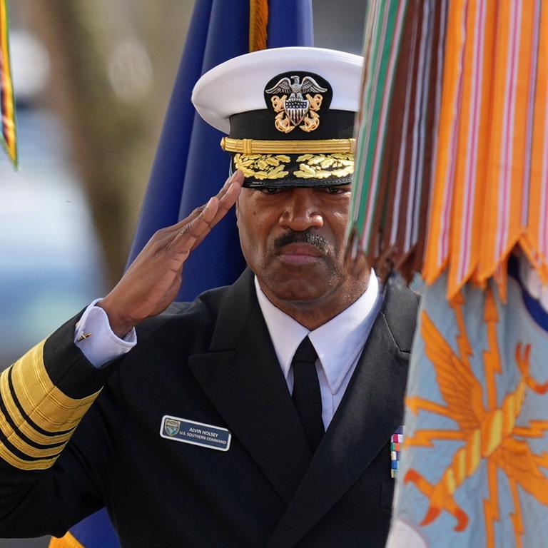 Das Bild zeigt Holseys Oberkörper in Paradeuniform. Er salutiert mit ernstem Blick. Neben ihm Fahnen von Armeeinheiten. 