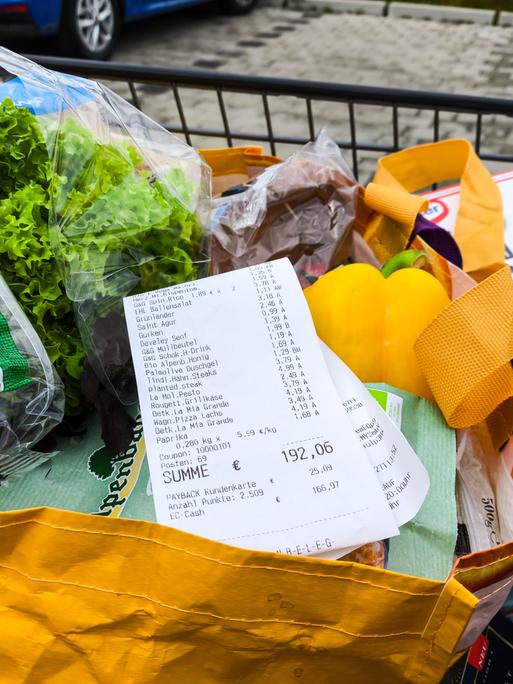 Ein Kassenbon mit einer Einkaufssumme von über 190 Euro liegt sichtbar auf frischen Lebensmitteln in einer Einkaufstasche, darunter Salat, Paprika und Tiefkühlpizza. 