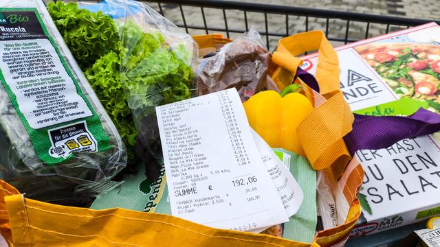 Ein Kassenbon mit einer Einkaufssumme von über 190 Euro liegt sichtbar auf frischen Lebensmitteln in einer Einkaufstasche, darunter Salat, Paprika und Tiefkühlpizza. 