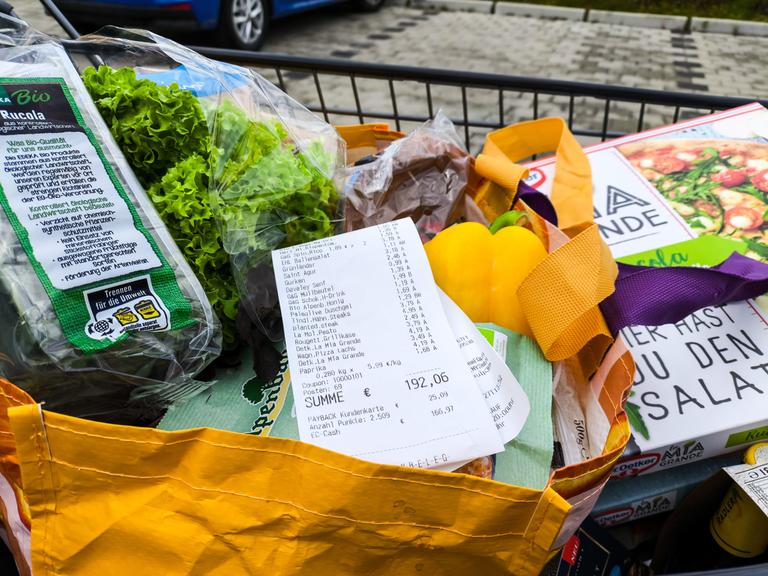 Ein Kassenbon mit einer Einkaufssumme von über 190 Euro liegt sichtbar auf frischen Lebensmitteln in einer Einkaufstasche, darunter Salat, Paprika und Tiefkühlpizza. 
