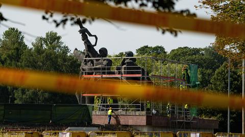 Hinter einem weißroten Absperrband sieht man das eingerüstete sowjetische Ehrenmal in Riga. Zwei Soldatenstatuen, die eine richtet ein Gewehr in die Luft.