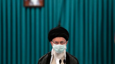 Der geistige Führer des iranischen Regimes Ayatollah Ali Khamenei mit einer Gesichtsmaske vor einem grünen Vorhang im Hintergrund. Auf dem Vorhang ein verschwommenes Bild von Ruhollah Khomeini.