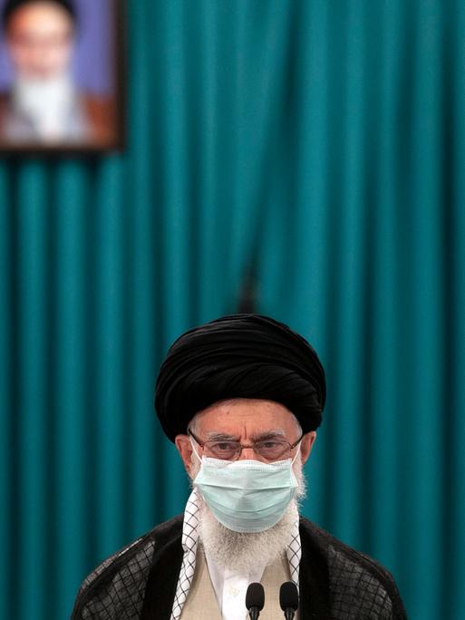 Der geistige Führer des iranischen Regimes Ayatollah Ali Khamenei mit einer Gesichtsmaske vor einem grünen Vorhang im Hintergrund. Auf dem Vorhang ein verschwommenes Bild von Ruhollah Khomeini.
