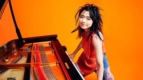 Eine Pianistin steht am Flügel, die Hände auf der Tastatur. Sie hat schwarze Haare und lächelt, alles ist sehr farbenfroh: orangefarbener Hintergrund, intensive Farben der Flügeltastatur.