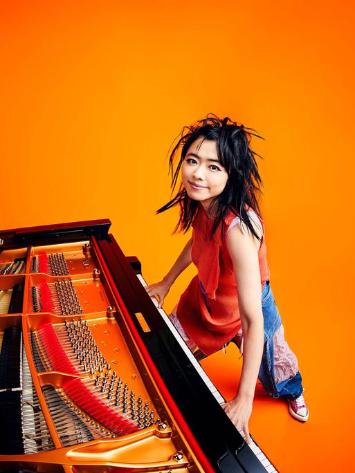 Eine Pianistin steht am Flügel, die Hände auf der Tastatur. Sie hat schwarze Haare und lächelt, alles ist sehr farbenfroh: orangefarbener Hintergrund, intensive Farben der Flügeltastatur.