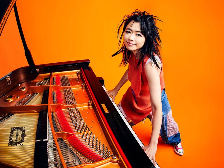 Eine Pianistin steht am Flügel, die Hände auf der Tastatur. Sie hat schwarze Haare und lächelt, alles ist sehr farbenfroh: orangefarbener Hintergrund, intensive Farben der Flügeltastatur.