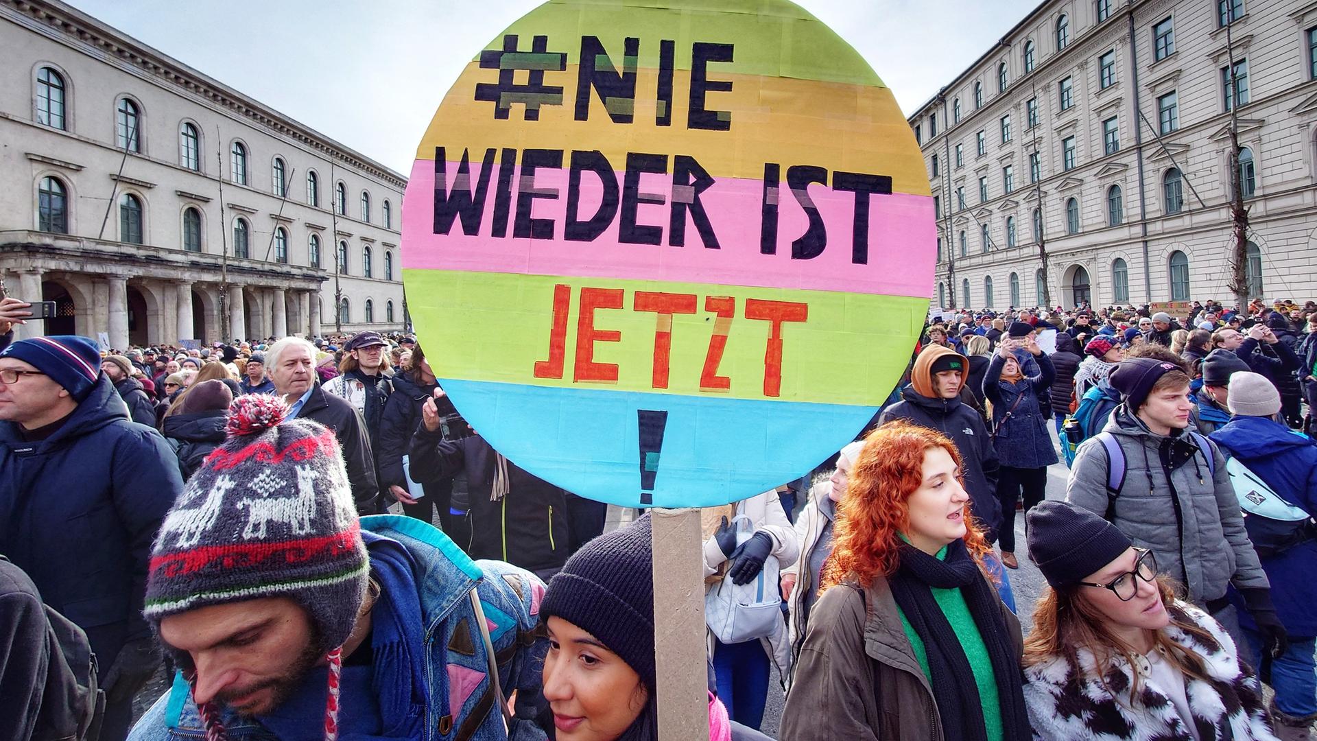 Eine Menschenmenge in einer Münchener Straße, im Vordergrund ein buntes rundes Plakat mit der Aufschrift "#Nie wieder ist jetzt"