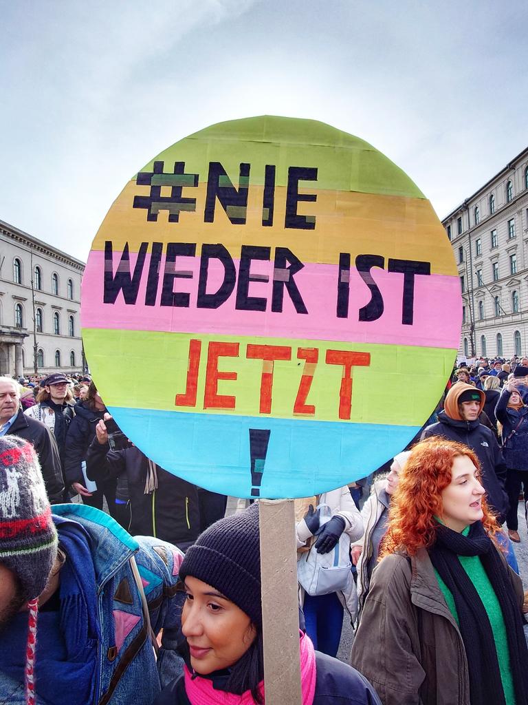 Eine Menschenmenge in einer Münchener Straße, im Vordergrund ein buntes rundes Plakat mit der Aufschrift "#Nie wieder ist jetzt" Eine Menschenmenge in einer Münchener Straße, im Vordergrund ein buntes rundes Plakat mit der Aufschrift "#Nie wieder ist jetzt"
