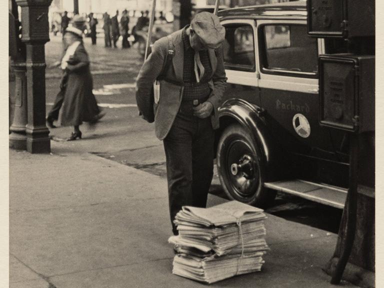 Unter der 6th Avenue steht ein Mann, der eine Werbetafel auf den Rücken geschnallt hat. Er beugt sich leicht nach vorne und liest die oberste Zeitung eines Zeitungsstapels.