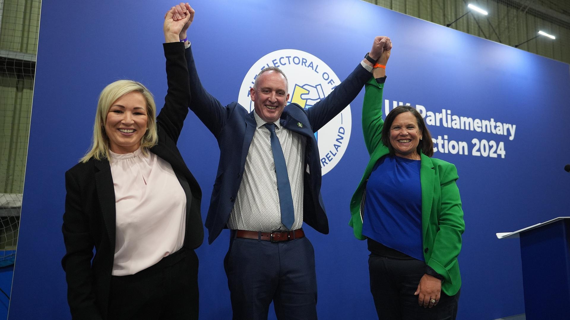 Die Sinn-Fein-Mitglieder Mallaghan, O'Neill und McDonald feiern ihr Ergebnis bei der Parlamentswahl. Sie stehen auf der Bühne und strecken die Arme hoch.