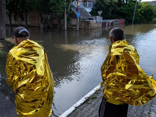 Bewohner von Cherson blicken auf die Überschwemmung in einer Straße. Sie sind in goldene Schutzfolien gehüllt, um sich zu wärmen. 