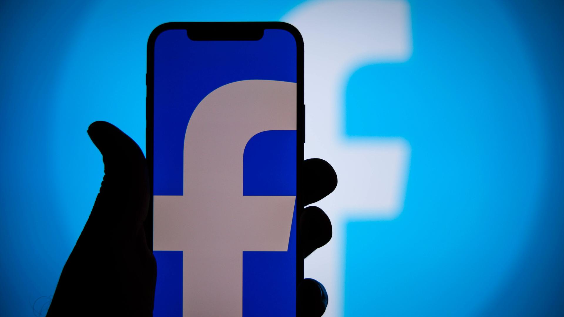 Eine Hand hält ein Smartphone, auf dessen blauem Bildschirm ein kleines weißes "f" zu sehen ist, das Symbol für Facebook.