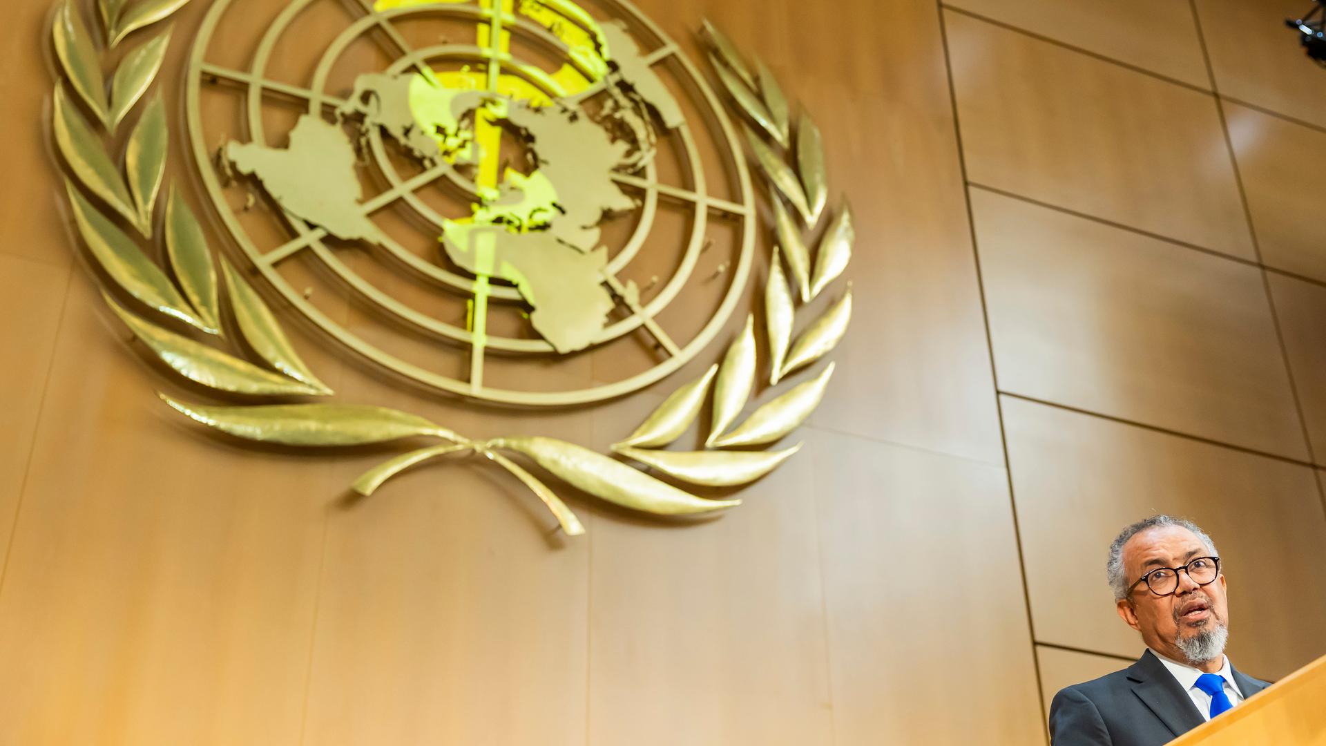 Tedros ist am unteren rechten Bildrand zu sehen, wie er an einem Rednepult spricht,. Hinter ihm groß eine holzvertäfelte Wanbd mit dem Wappen der WHO. 