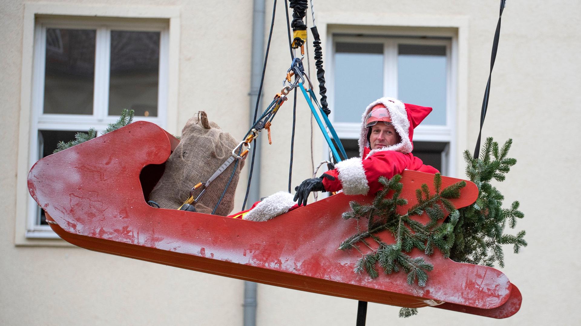 Ein großer roter Schlitten, in dem ein als Nikolaus verkleideter Mann sitzt, schwebt vor einem Haus in der Luft, gehalten von einem starken Zugseil.