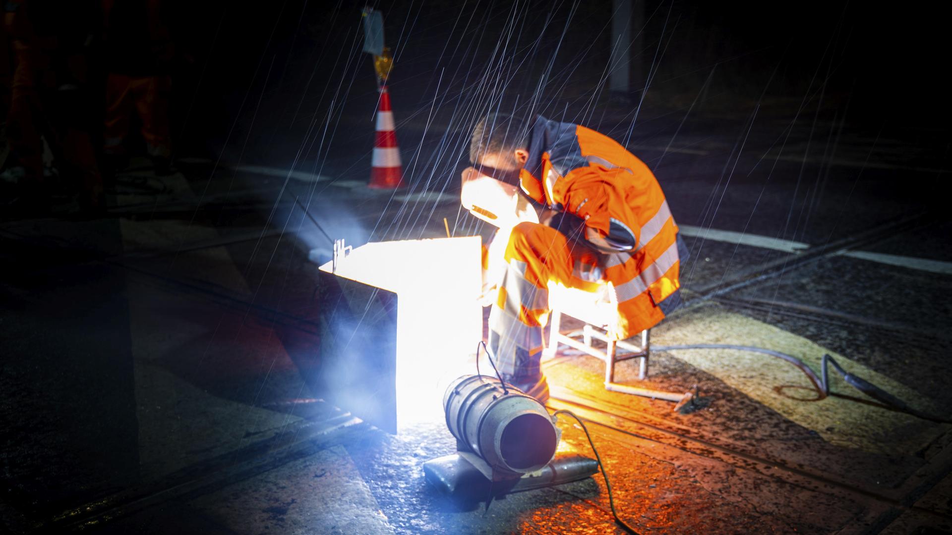 Ein Mitarbeiter eines Bautrupps schweißt nachts bei Regen verschlissene Straßenbahngleise mit einem Gesichtsschutz. 