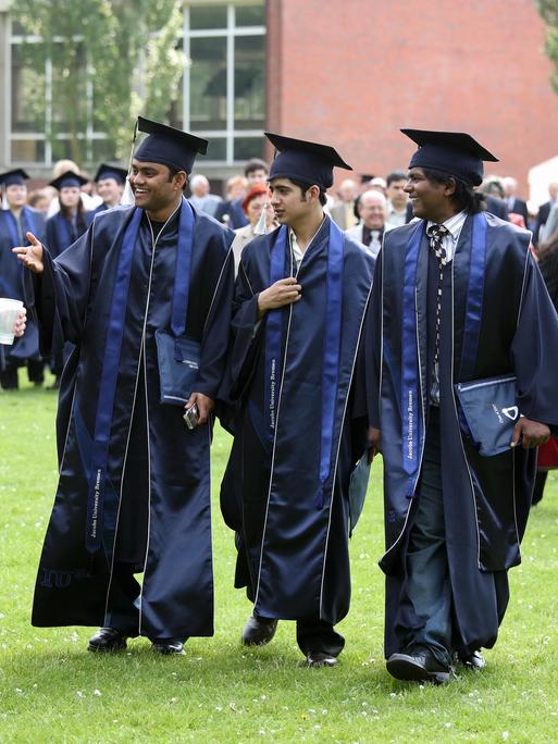 Abschlussfeier an der Uni in Bremen. Junge Menschen mit Doktorhüten gehen fröhlich über eine Wiese.