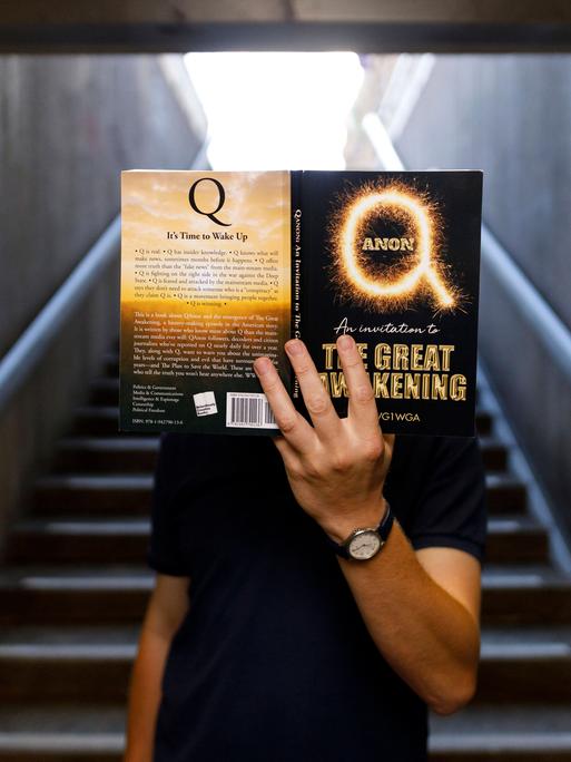 Jemand verdeckt seinen Kopf mit dem Amazon-Bestseller Qanon - An Invitation to the Great Awakening . Köln, 05.07.2020 *