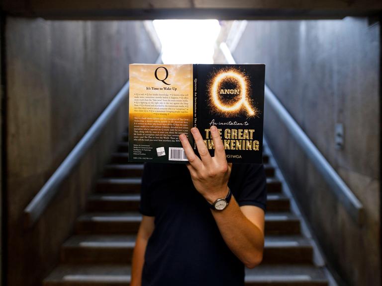 Jemand verdeckt seinen Kopf mit dem Amazon-Bestseller Qanon - An Invitation to the Great Awakening . Köln, 05.07.2020 *