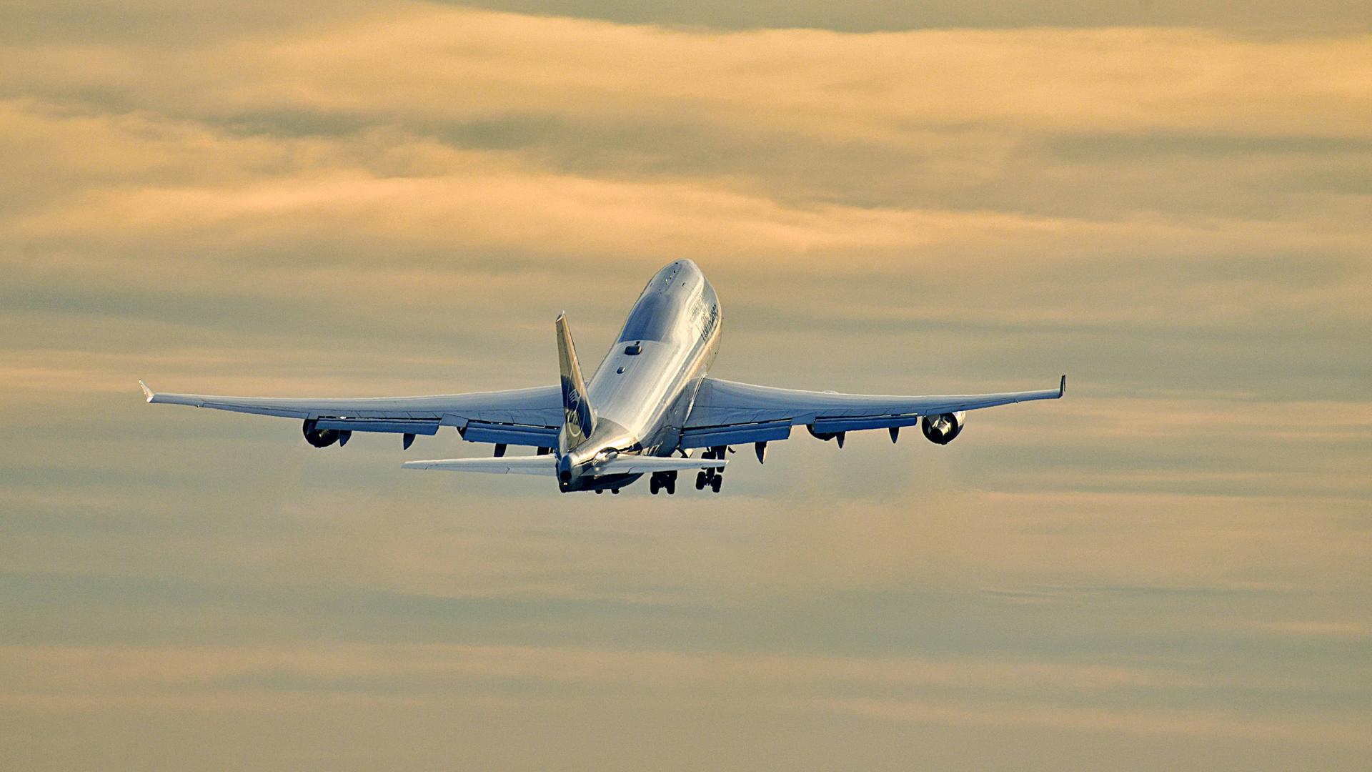 Foto einer startende Boeing 747 der Lufthansa am Frankfurter Flughafen, die vor einem orangefarbenen, bewölkten Abendhimmel aufsteigt.