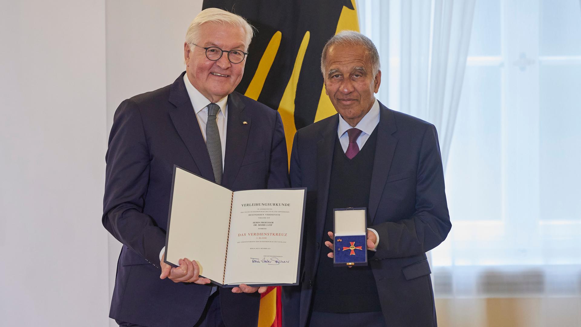 Bundespräsident Frank-Walter Steinmeier hält eine Urkunde in der Hand, neben ihm steht Mojib Larif mit dem Verdienstorden in einem Kästchen.