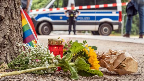 Passanten haben 80 Meter vom Tatort des CSD-Anschlags eine Regenbogenfahne, ein Grablicht und Sonnenblumen niedergelegt. Im Hintergrund ist ein Polizeibus zu erkennen.