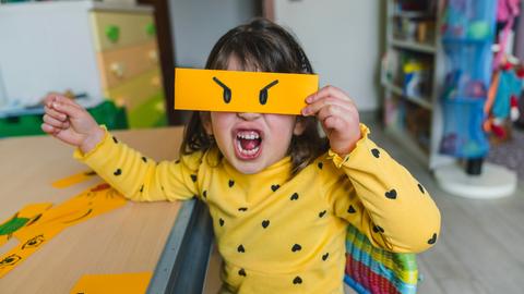 Ein Mädchen macht ein wütendes Gesicht und hält sich die auf Papier dazu passend gemalten, wütenden Augen vors Gesicht.