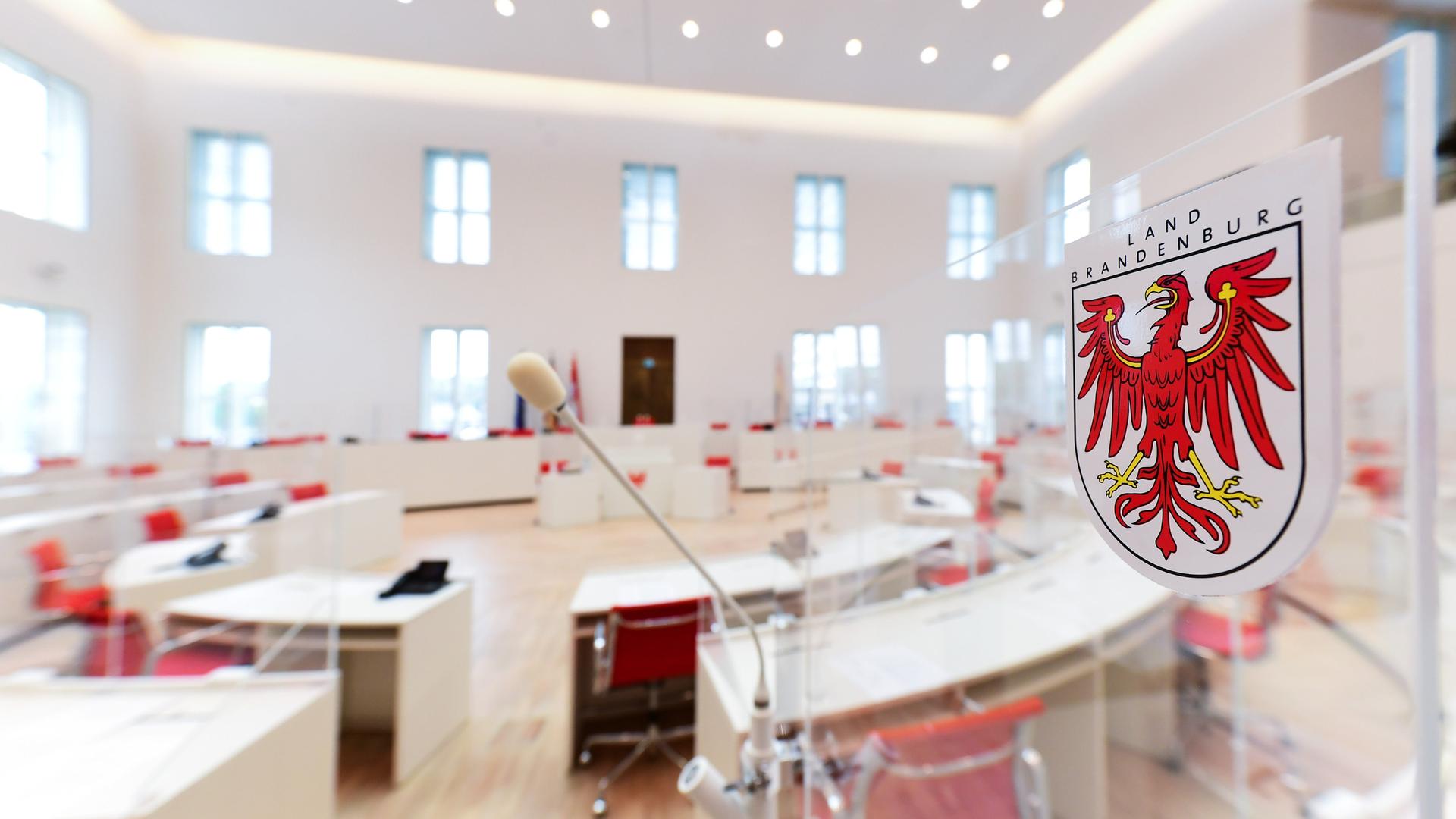 Blick in den Plenarsaal des Brandenburger Landtages, im Vordergrund ein Mikrofon, rechts das Wappen des Landes Brandenburg