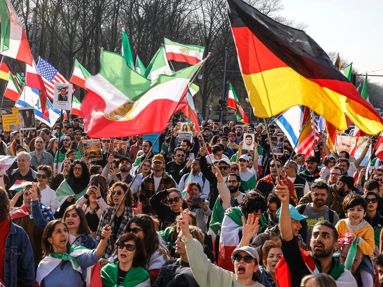 Das Foto zeigt eine Menschenmenge, die iranische, deutsche, US-amerikanische und israelische Flaggen schwenkt und gegen das iranische Regime protestiert. Die Demonstration fand am 7. März 2026 in Berlin statt.