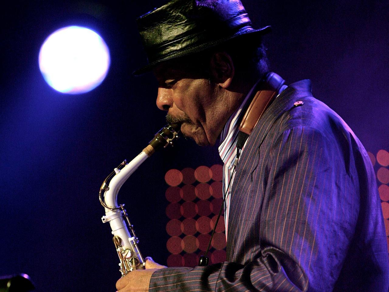 Man sieht Saxophonisten Ornette Coleman im Profil, wie er 2015 mit schwarzem Hut auf der Bühne sein Instrument vor einem Mikrofon spielt.