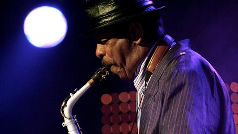Man sieht Saxophonisten Ornette Coleman im Profil, wie er 2015 mit schwarzem Hut auf der Bühne sein Instrument vor einem Mikrofon spielt.