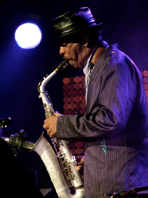 Man sieht Saxophonisten Ornette Coleman im Profil, wie er 2015 mit schwarzem Hut auf der Bühne sein Instrument vor einem Mikrofon spielt.