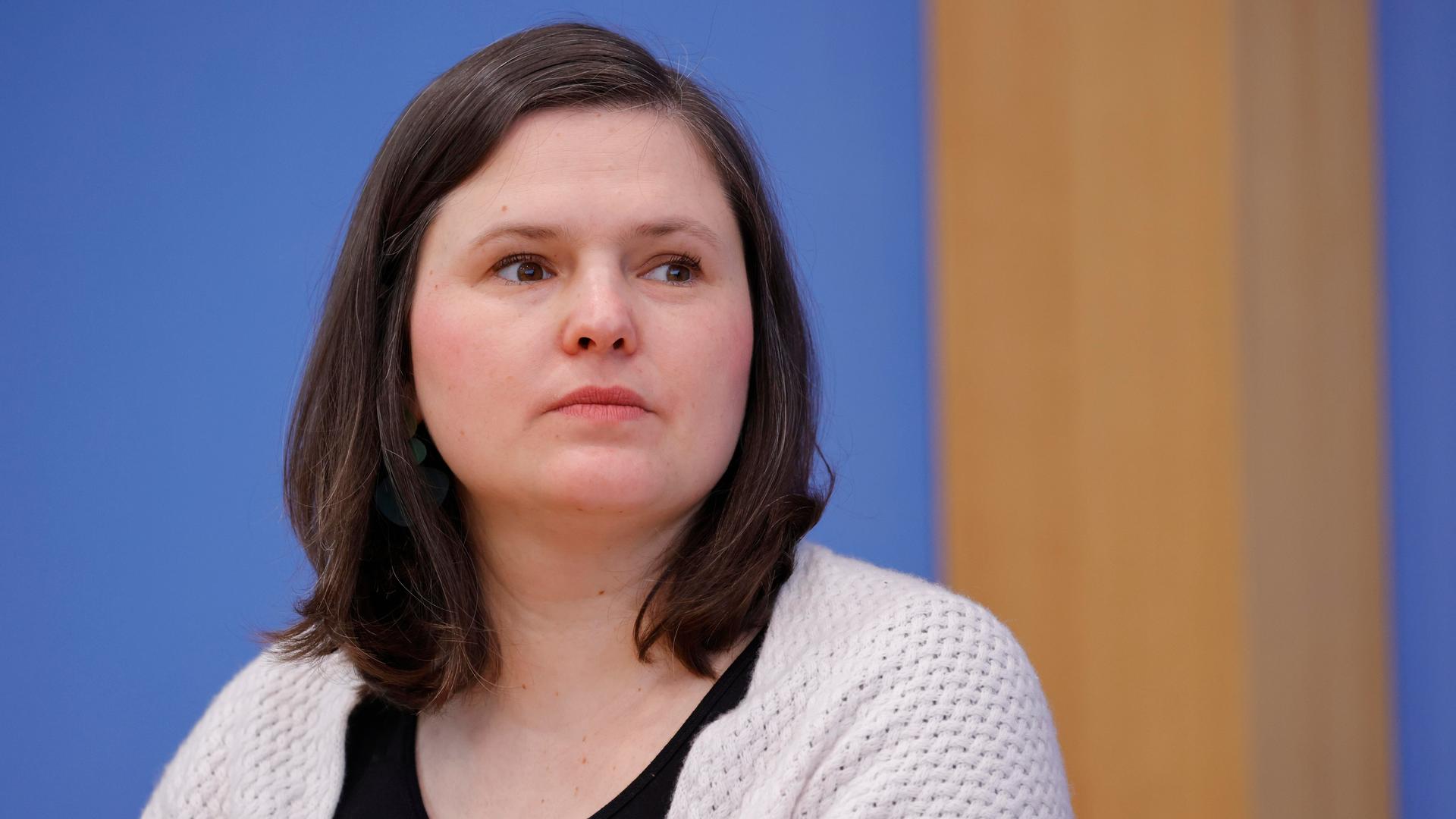 Sara Nanni, MdB Bündnis 90/Die Grünen bei einer Bundespressekonferenz. Sie trägt eine beige Strickjacke über einem schwarzen T-Shirt und schaut nachdenklich und mit ernstem Blick aus dem Bild heraus.