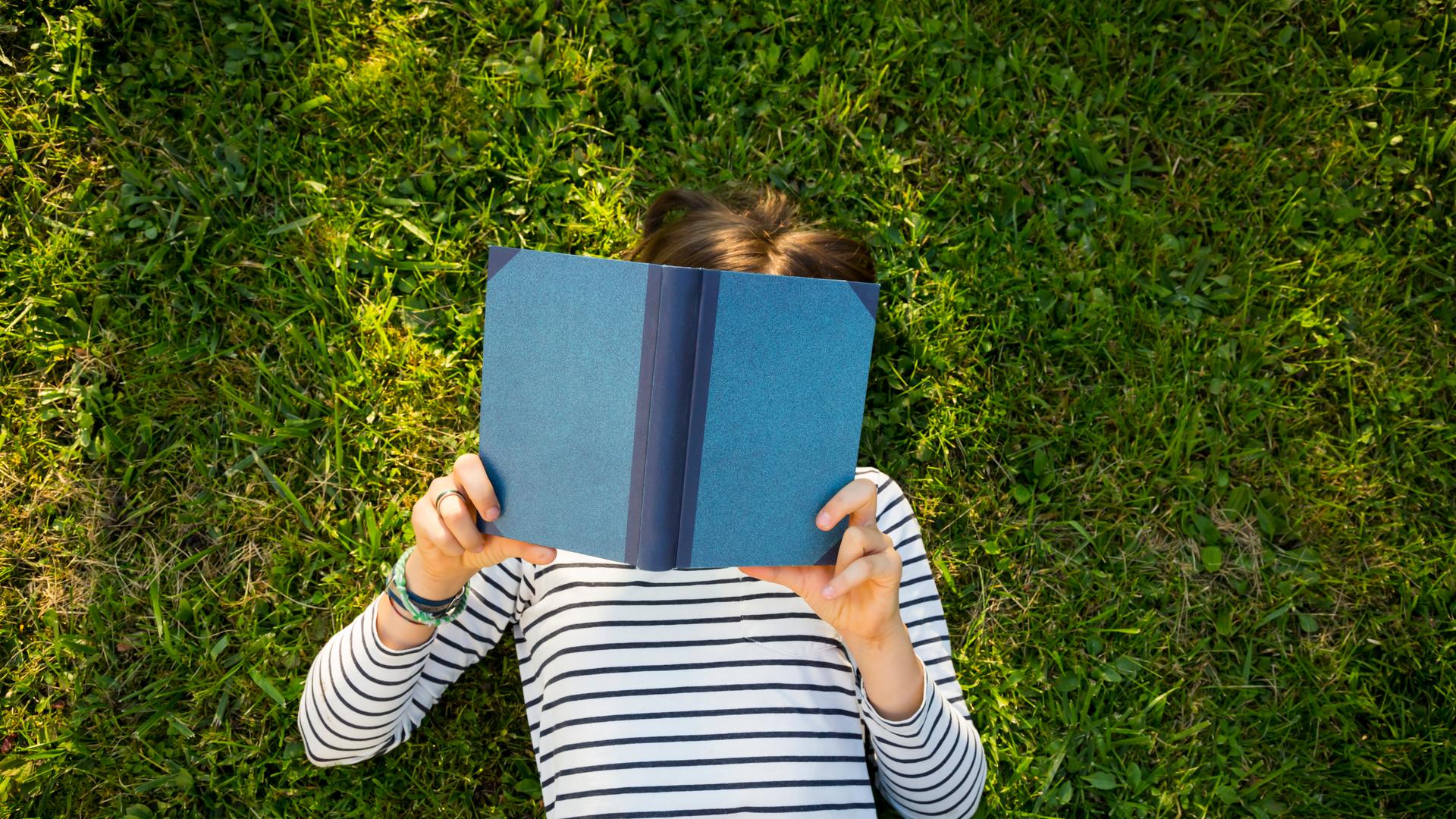 Ein Mädchen liegt auf einer Wiese und liest ein Buch. Ansicht von oben. Das Gesicht des Kindes ist unter dem Buch nicht zu erkennen.