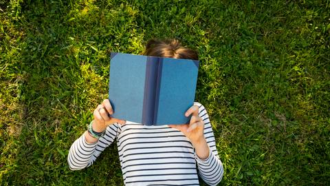 Ein Mädchen liegt auf einer Wiese und liest ein Buch. Ansicht von oben. Das Gesicht des Kindes ist unter dem Buch nicht zu erkennen.