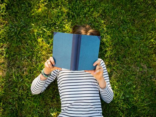 Ein Mädchen liegt auf einer Wiese und liest ein Buch. Ansicht von oben. Das Gesicht des Kindes ist unter dem Buch nicht zu erkennen.